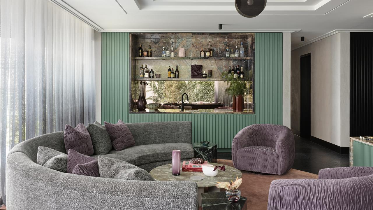 A modern living room featuring a curved gray fabric sofa with purple throw pillows, a round coffee table, a matching purple armchair, and a green ribbed accent wall behind a stone shelf display.