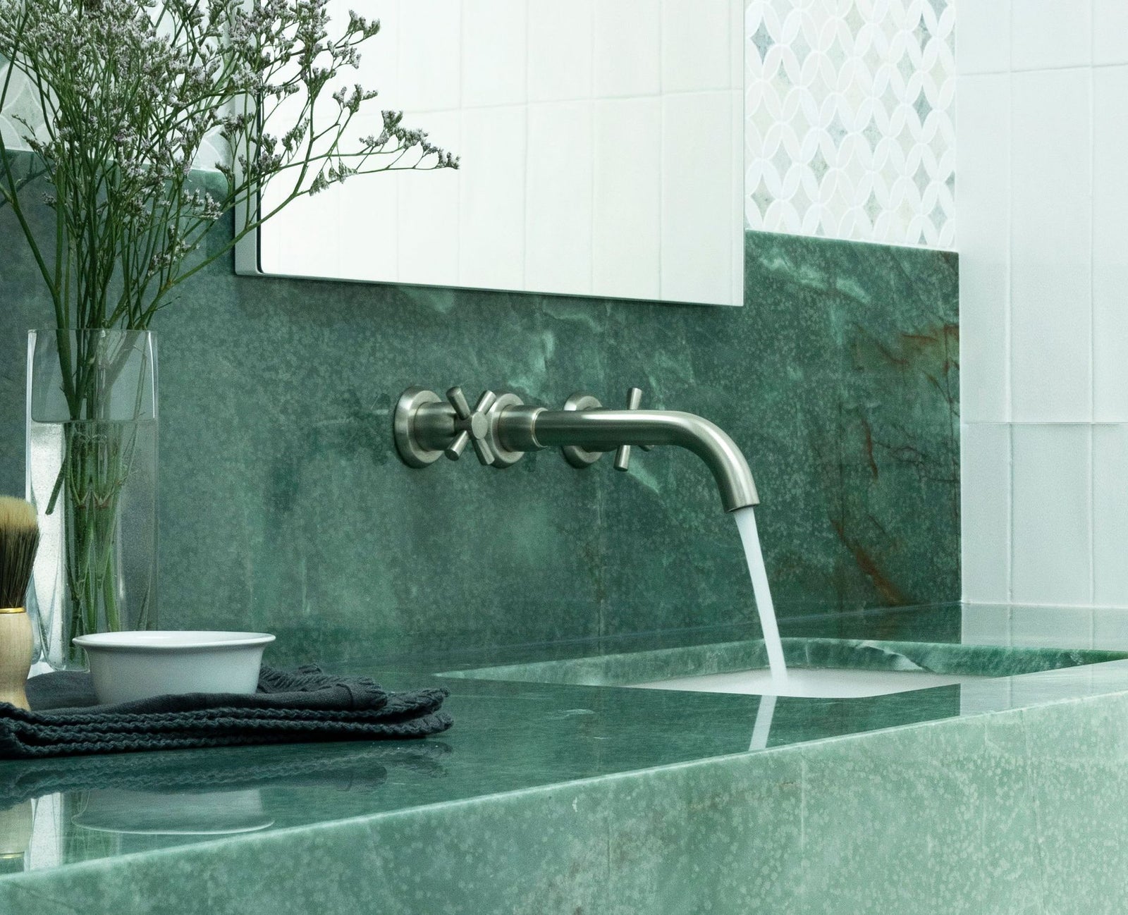 A modern wall-mounted sink faucet with a curved spout and cross handles, shown with water running into a green stone basin. The sink is set against a matching green marble backsplash with white tiles and a mirror in the background.